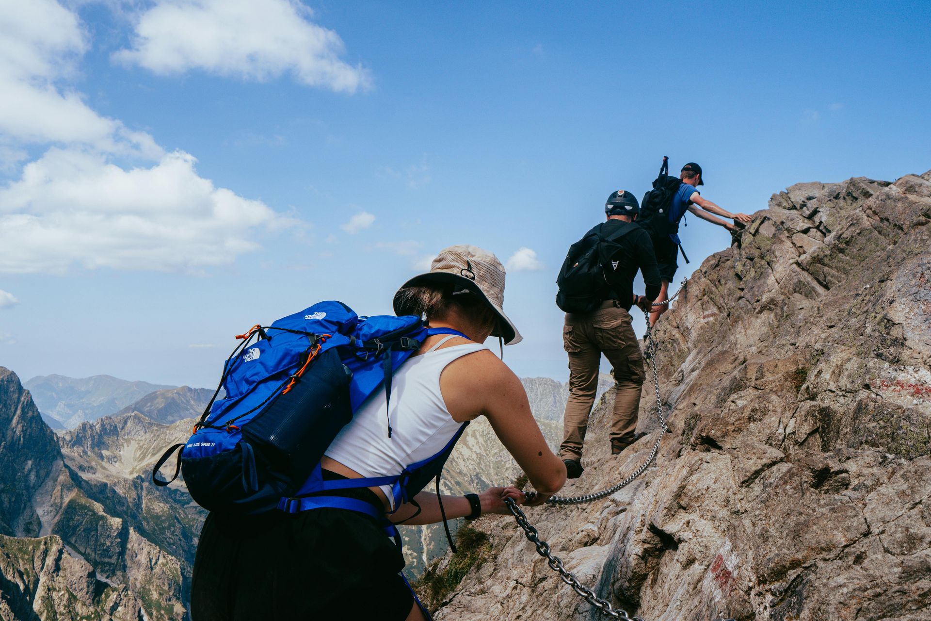 Three people on a steep mountainside, using a chain attached to the rock to support them while climbing.