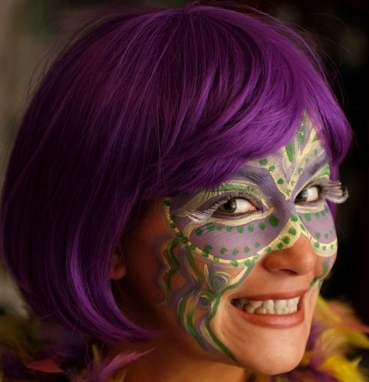 A smiling person wearing a purple bob wig and intricate face paint in purple, green, and gold, featuring a decorative masquerade mask design around the eyes.