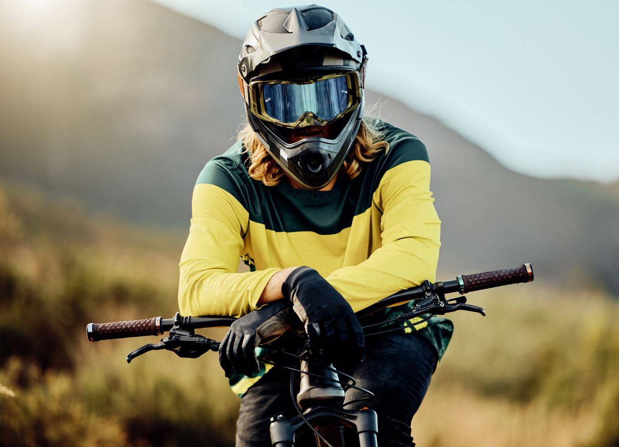 A mountain biker in a full-face helmet with mirrored goggles and a green and yellow long-sleeve jersey leans over the handlebars of their bike, with a blurred mountain landscape in the background.