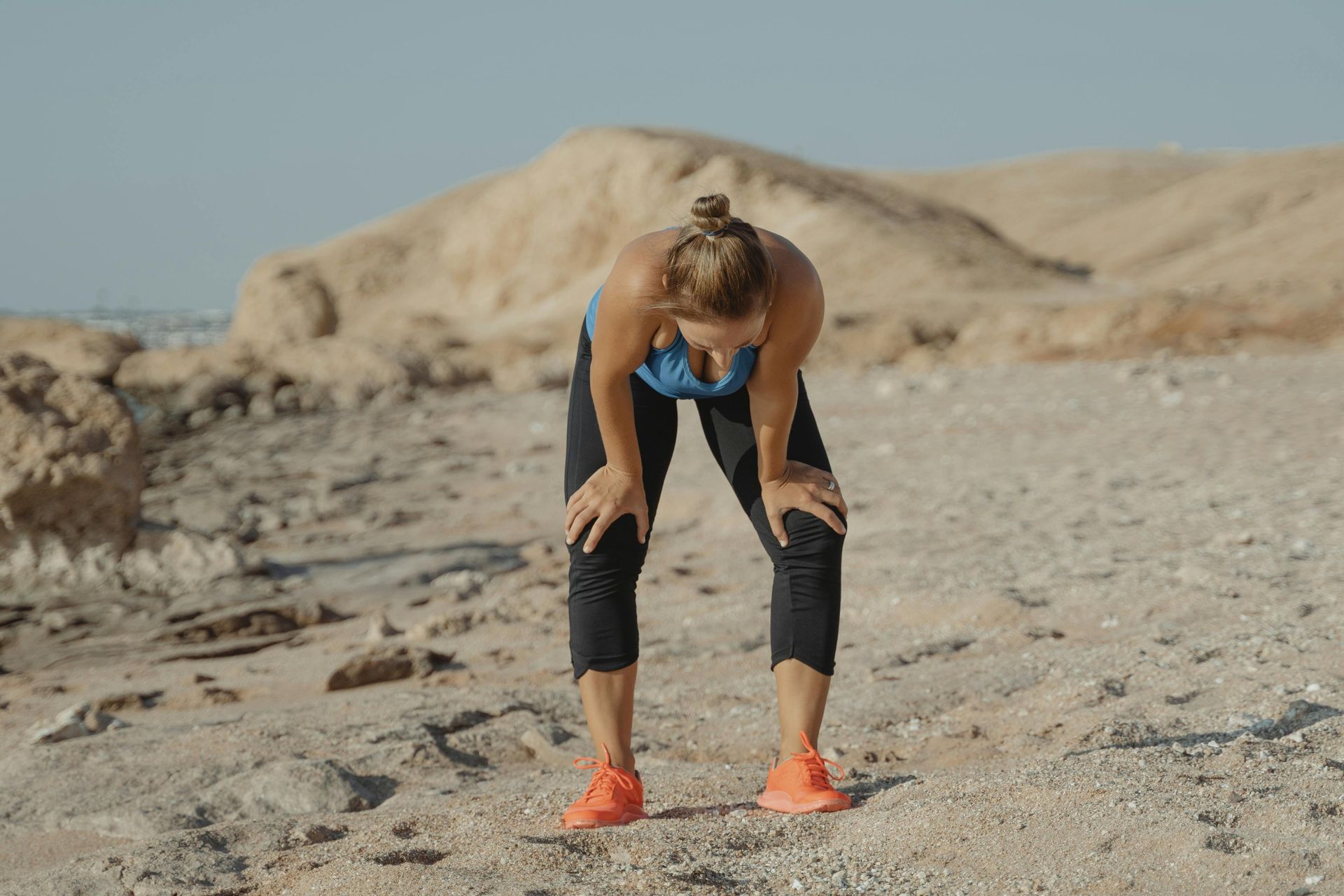 Woman in athletic gear bent over with hands on knees on a rocky landscape, looking spent