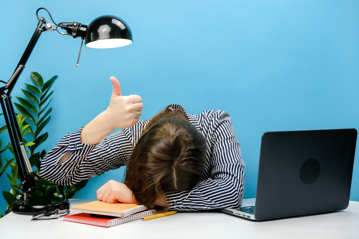 A person in a striped shirt slumped face-down on a desk covered with notebooks, giving a thumbs-up with one hand while a laptop and desk lamp sit nearby, against a bright blue background.