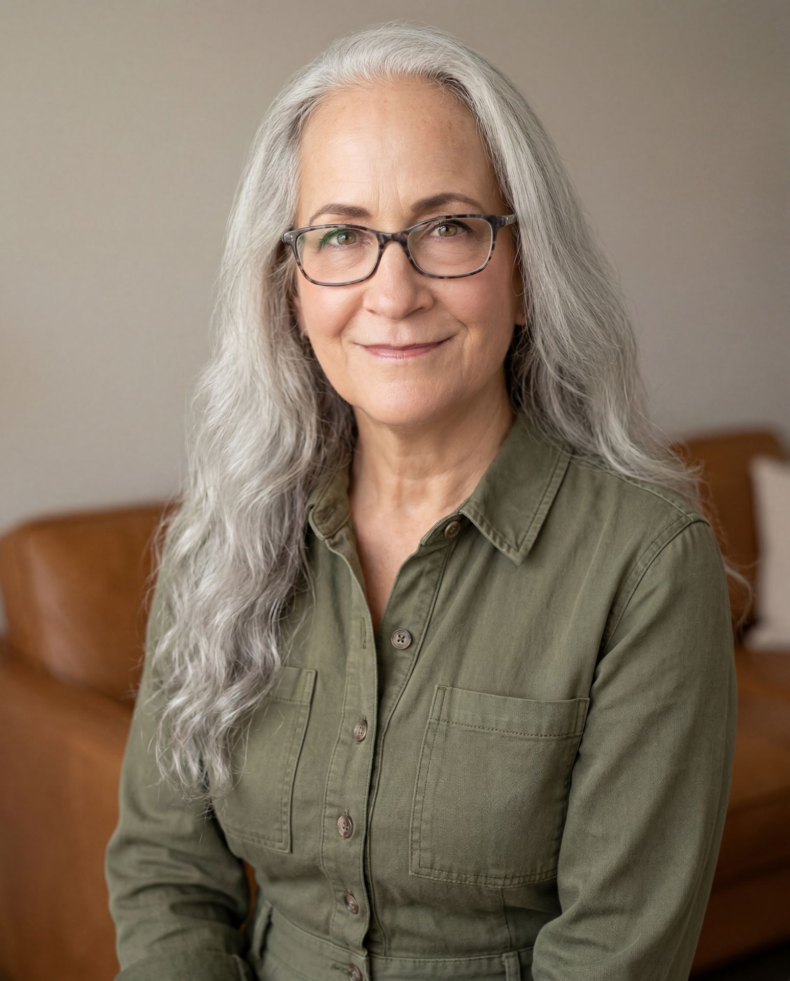 A warm, experienced psychotherapist with long gray hair and glasses smiles gently in a cozy home office, reflecting the calm, steady presence she offers parents of anxious children through online parent therapy in California.