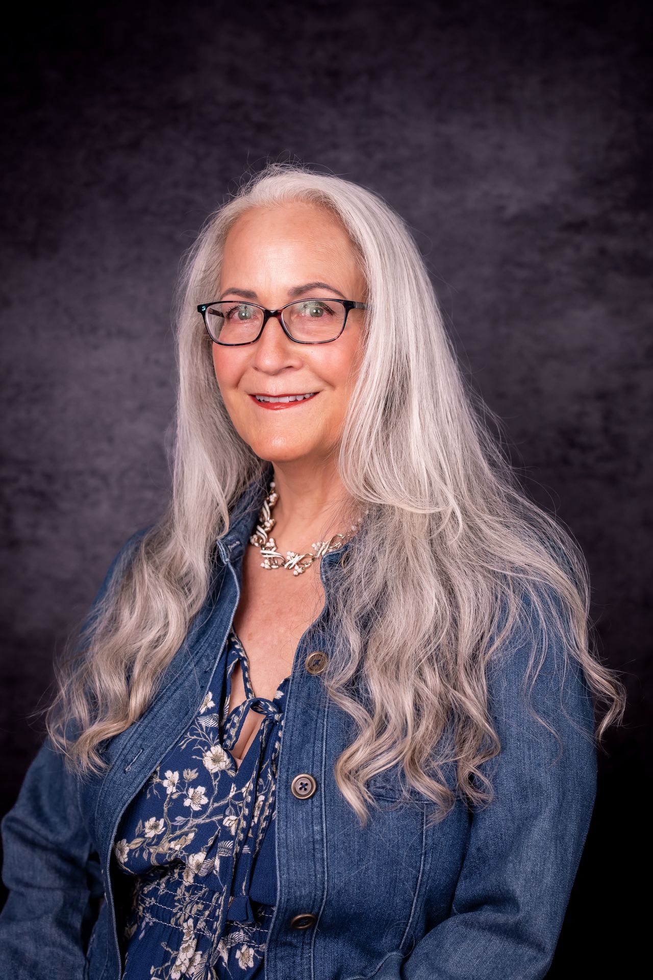 Abby McCarrel, a warm and experienced psychotherapist with long silver hair, glasses, and a gentle smile, sits on an outdoor patio in a blue floral dress and jean jacket. Her calm, compassionate presence reflects her work providing online family therapy, helping parents and children strengthen connection, navigate challenges, and build emotional safety through trauma-informed, brain-based support.