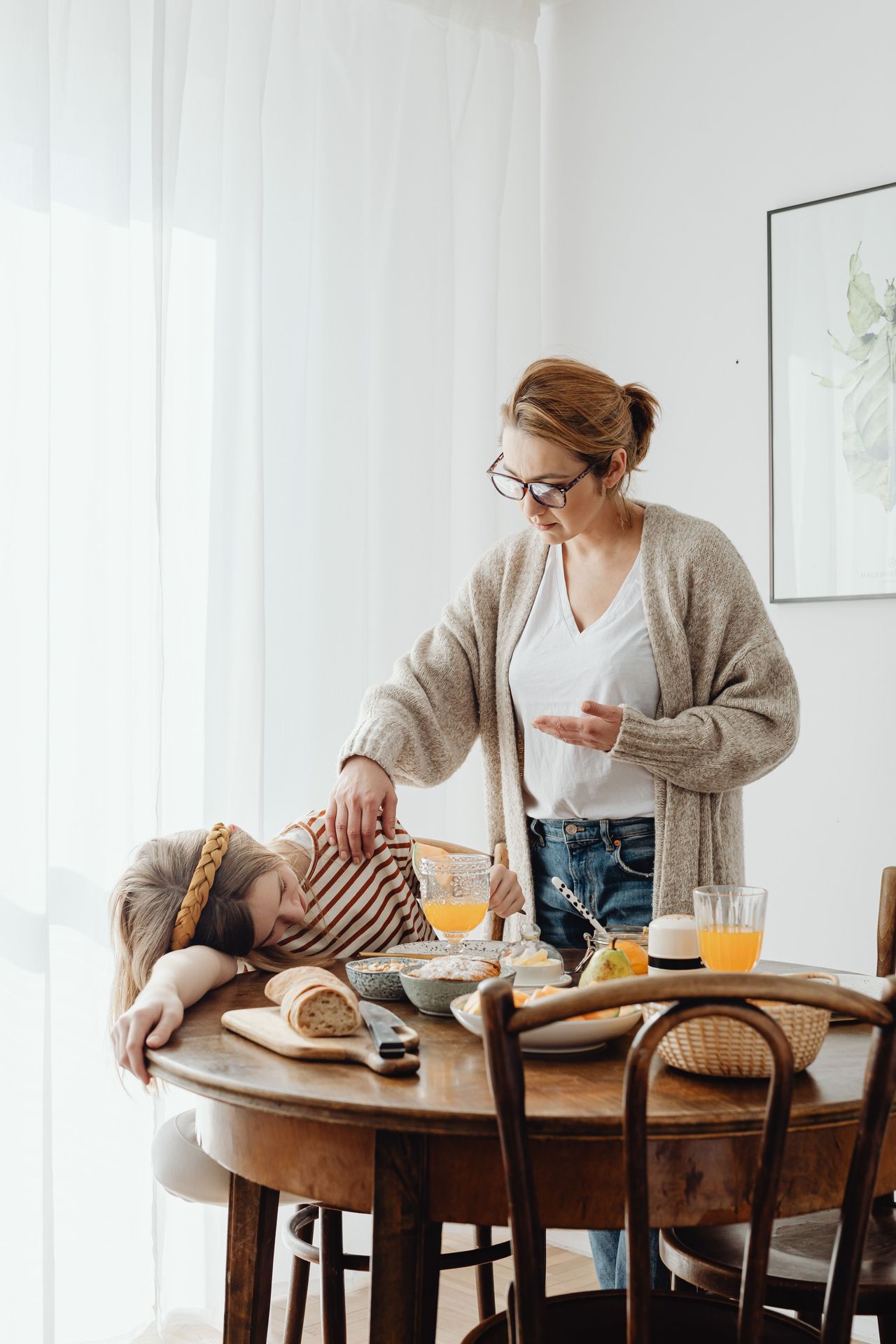 A concerned mother in glasses gently checks on her child, who is slumped over a breakfast table, capturing the quiet exhaustion of parenting a child with big, baffling behaviors and the daily toll it takes on the whole family.