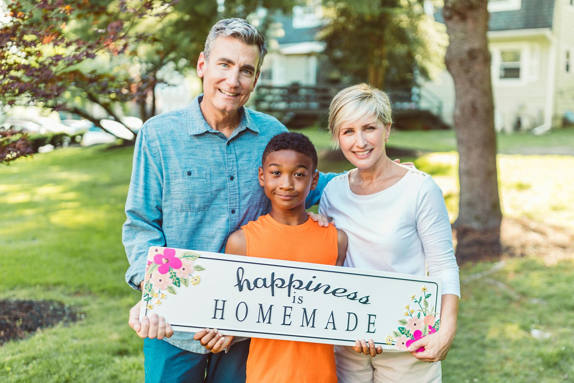 A joyful white couple stands with their Black son outside his new home, holding a sign that reads 'Happiness is Homemade.' This heartwarming moment captures the support and connection nurtured through adoption services, helping families build lasting bonds and create loving, stable homes for adopted children.