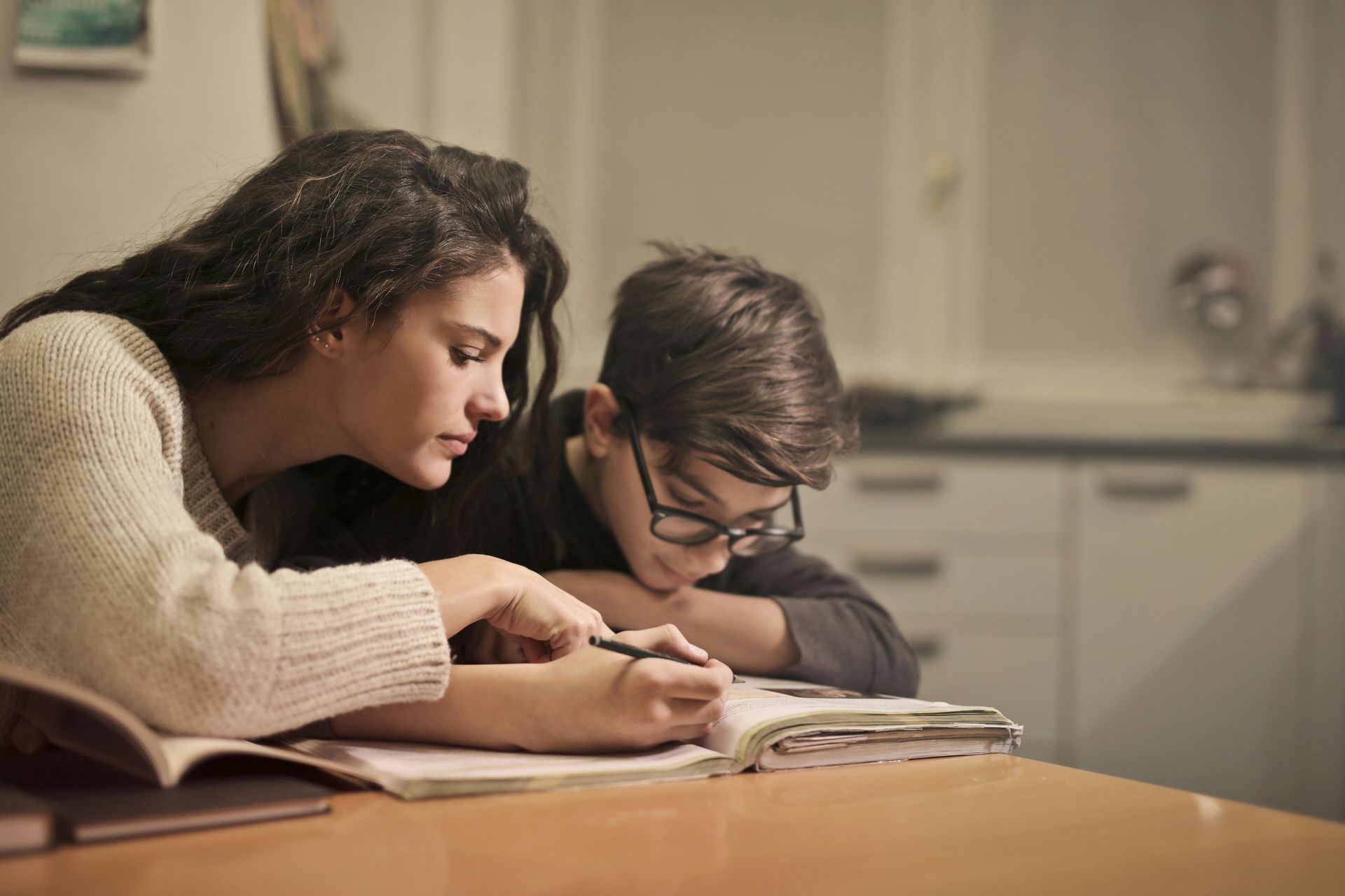 Family counseling for 2e support at home. A mother and her twice-exceptional (2e) son sit at the kitchen table, focused on a workbook. The boy, wearing glasses, leans in as they work together, highlighting the importance of family guidance and educational support for gifted and neurodivergent children
