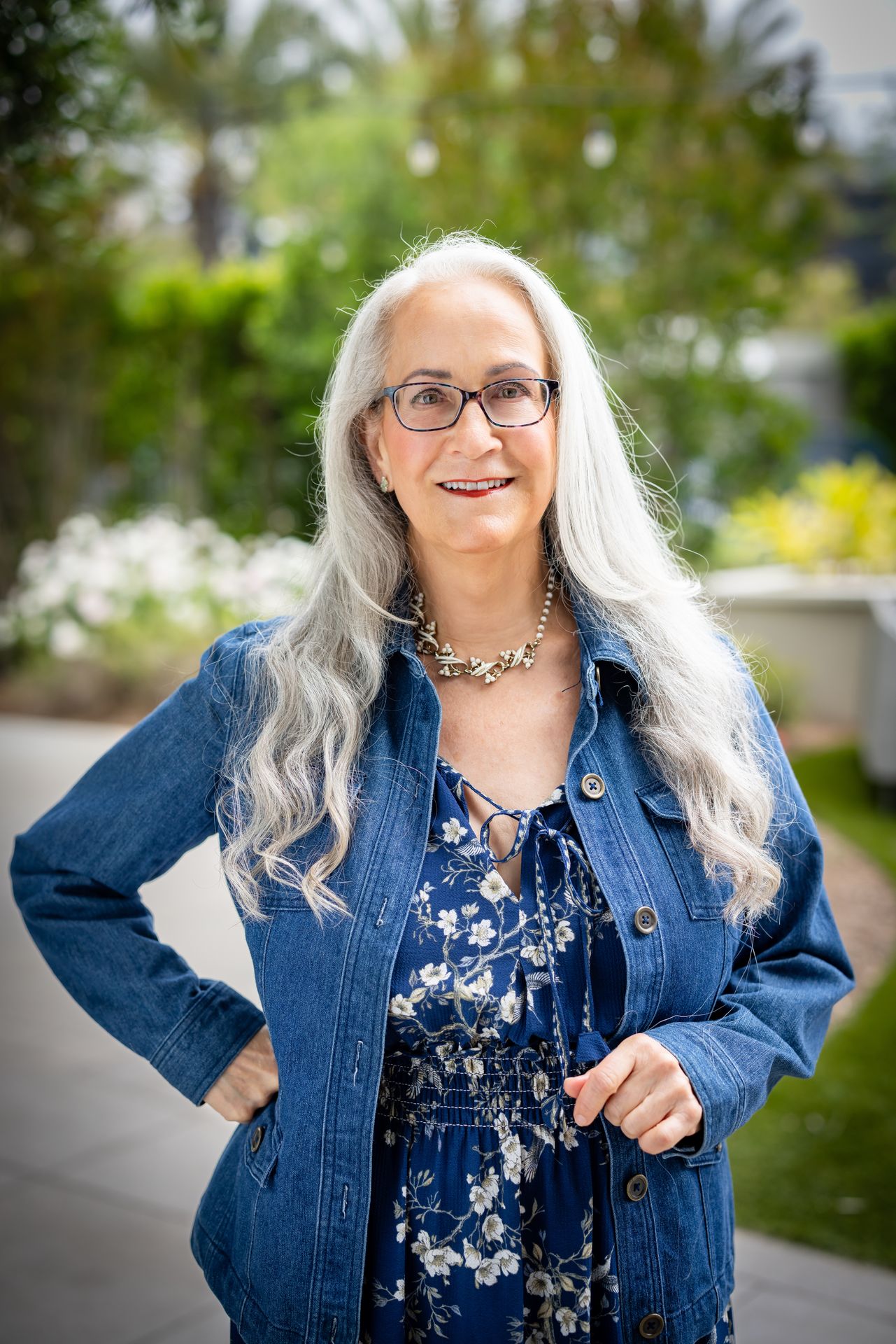 Abby McCarrel, a warm and experienced psychotherapist with long silver hair, glasses, and a gentle smile, sits on an outdoor patio in a blue floral dress and jean jacket. Her compassionate presence reflects her work in therapy for extended family relationships, helping families navigate conflicts, strengthen bonds, and create more safety and connection across generations.