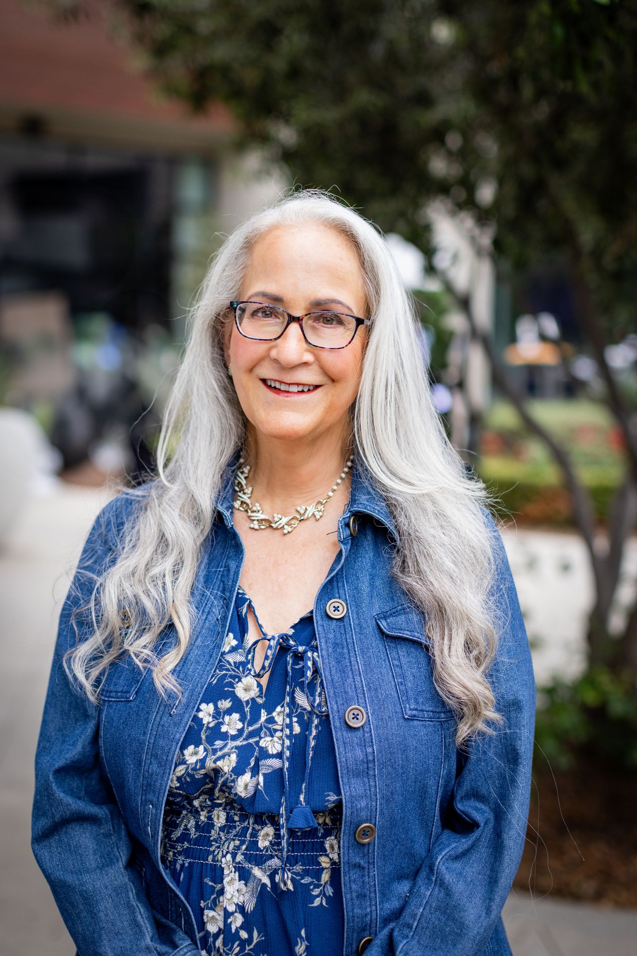 Abby McCarrel, a warm and experienced psychotherapist with long silver hair, glasses, and a gentle smile, sits on an outdoor patio in a blue floral dress and jean jacket. Her grounded presence reflects her work supporting adults diagnosed late with ADHD, offering compassionate, brain-based therapy to help them make sense of their experiences, reduce shame, and create meaningful change in their lives.