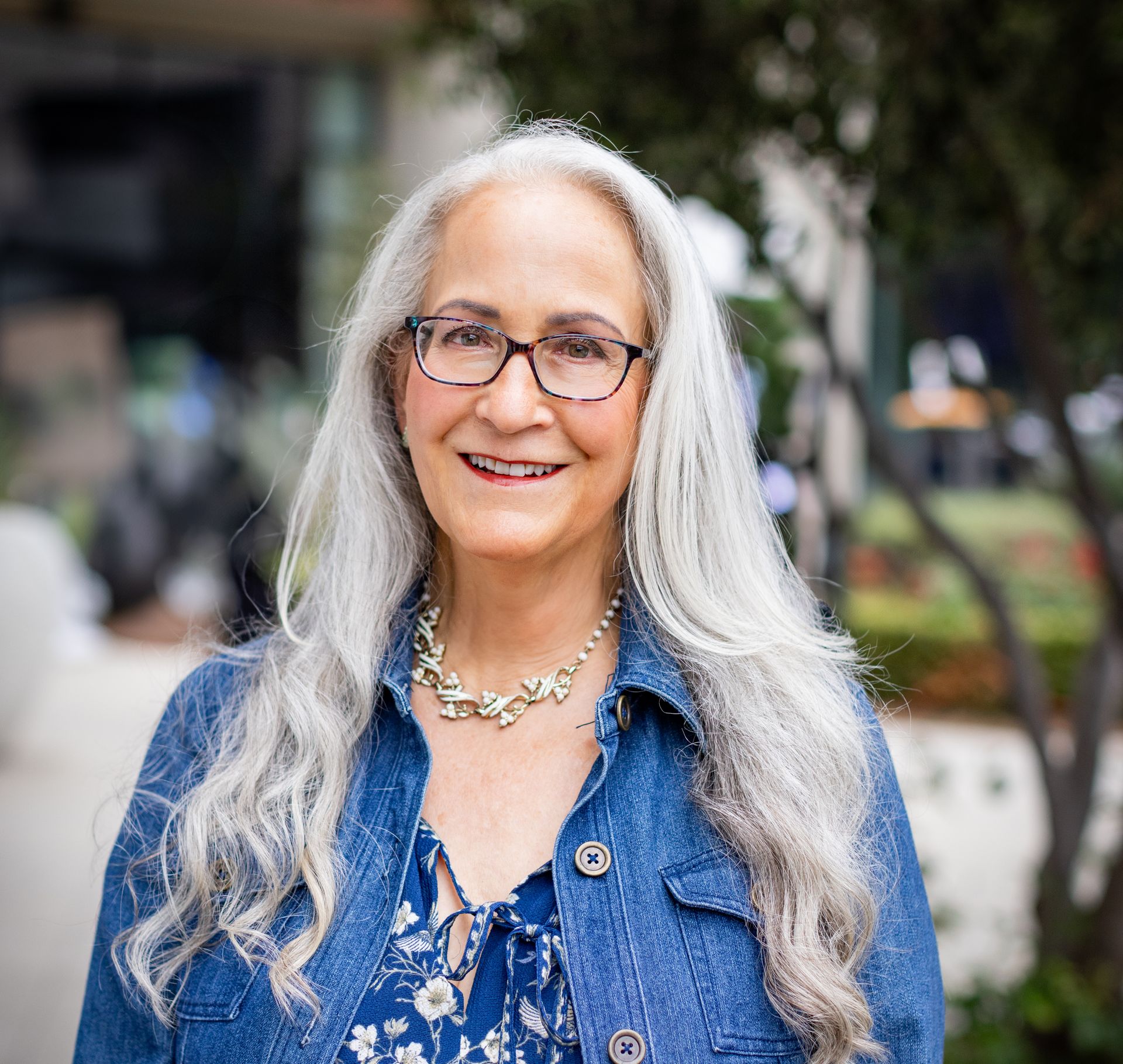 Abby McCarrel, a warm and experienced psychotherapist with long silver hair, glasses, and a gentle smile, sits on an outdoor patio wearing a blue floral dress and jean jacket. Her calm, thoughtful presence reflects her work in parent coaching grounded in brain science, helping parents understand their child’s nervous system, reduce stress, and strengthen connection through trauma-informed, neuroscience-based support.