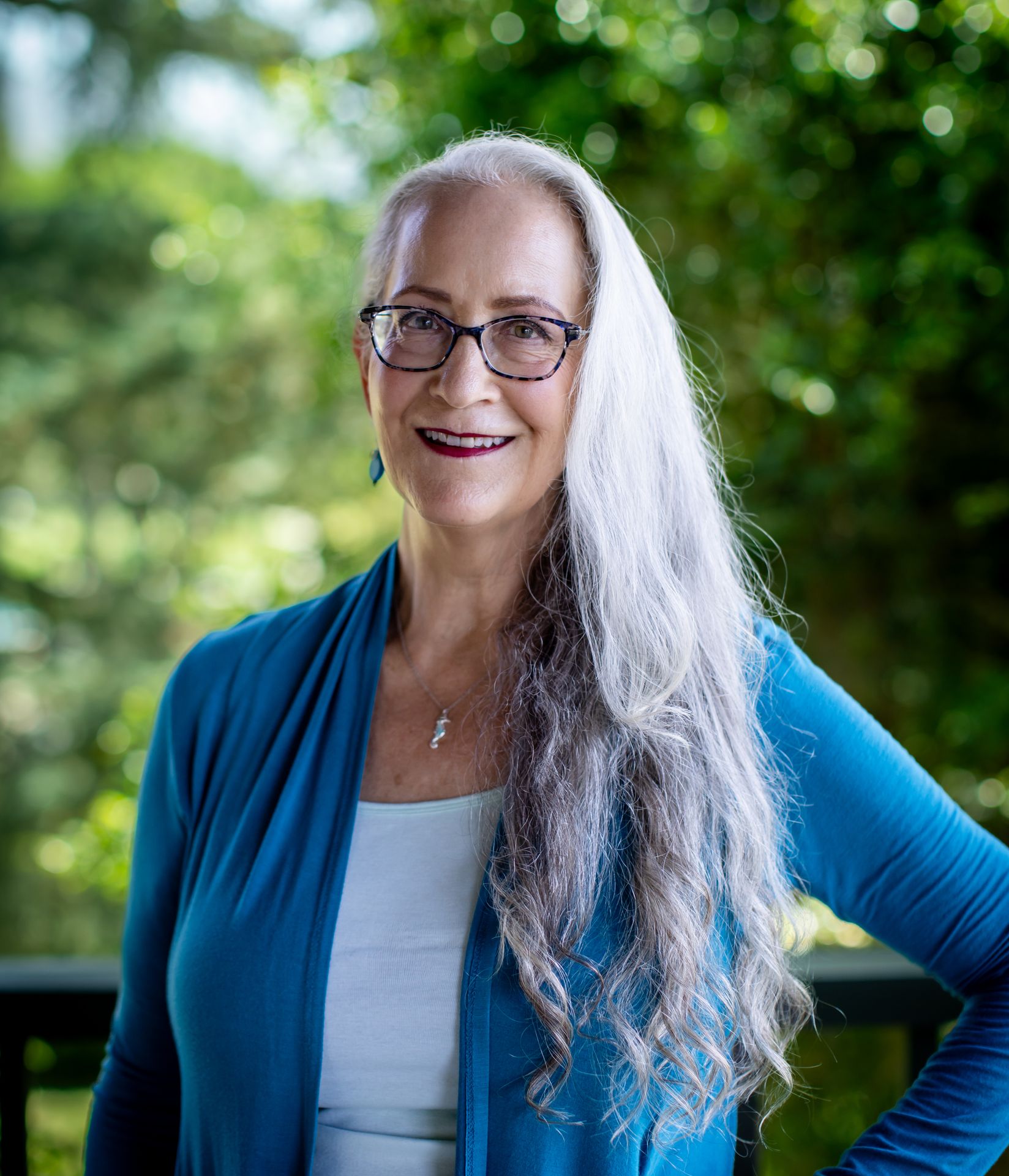 Abby McCarrel, a warm and experienced psychotherapist with long white hair and glasses, stands outdoors with a confident smile and hand on her hip, surrounded by lush greenery. Her calm, grounded presence reflects what families can expect from online family therapy in California—a trauma-informed, brain-based approach that helps parents feel supported as they rebuild connection, reduce conflict, and foster healing at home.