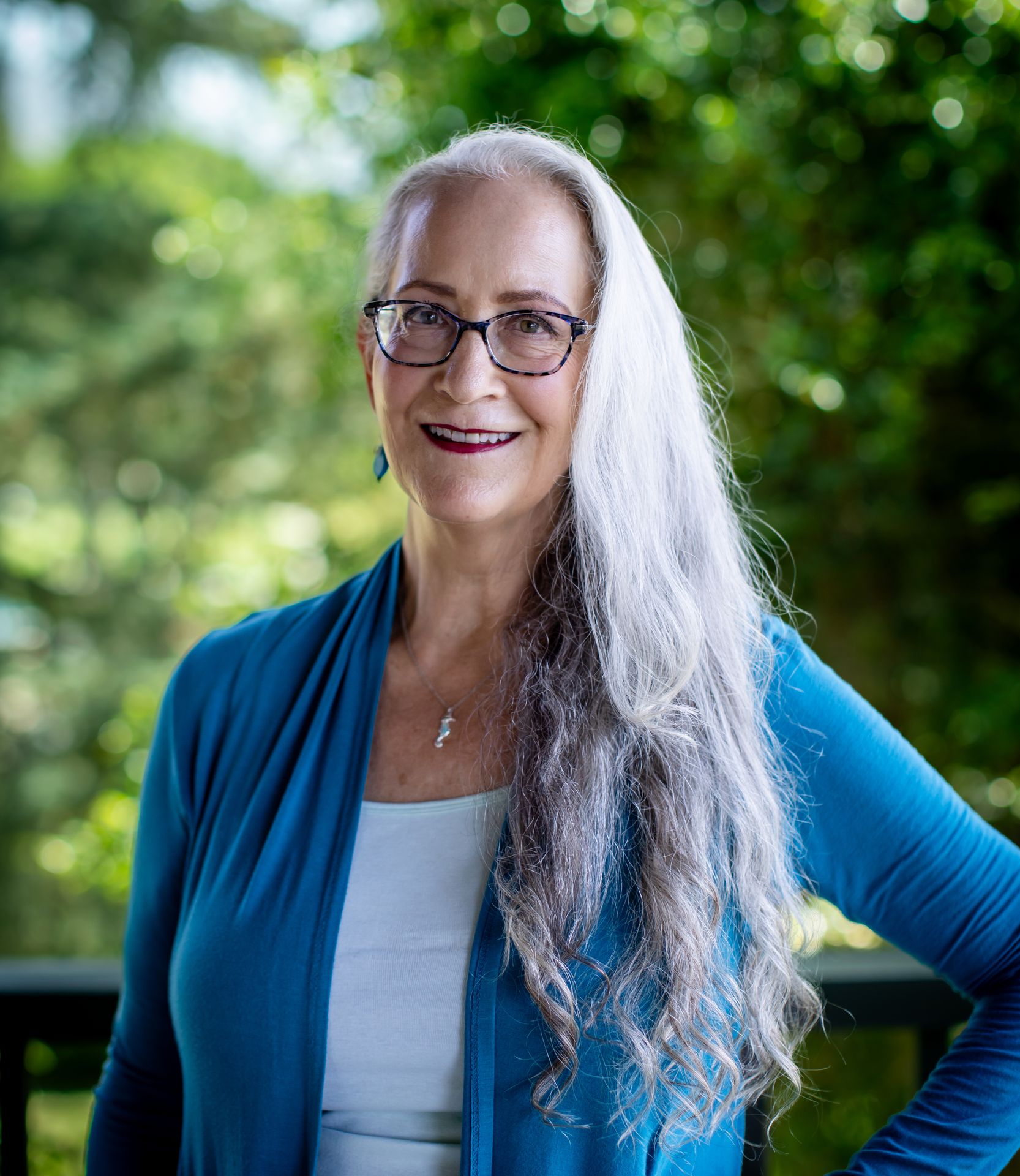 Abby McCarrel, a warm and experienced psychotherapist with long white hair and glasses, stands outdoors with her hand on her hip, surrounded by lush greenery. Her calm, confident presence reflects her work providing counseling for parents of ADHD children, offering brain-based, trauma-informed support to help families reduce overwhelm, improve connection, and respond with greater understanding and compassion.