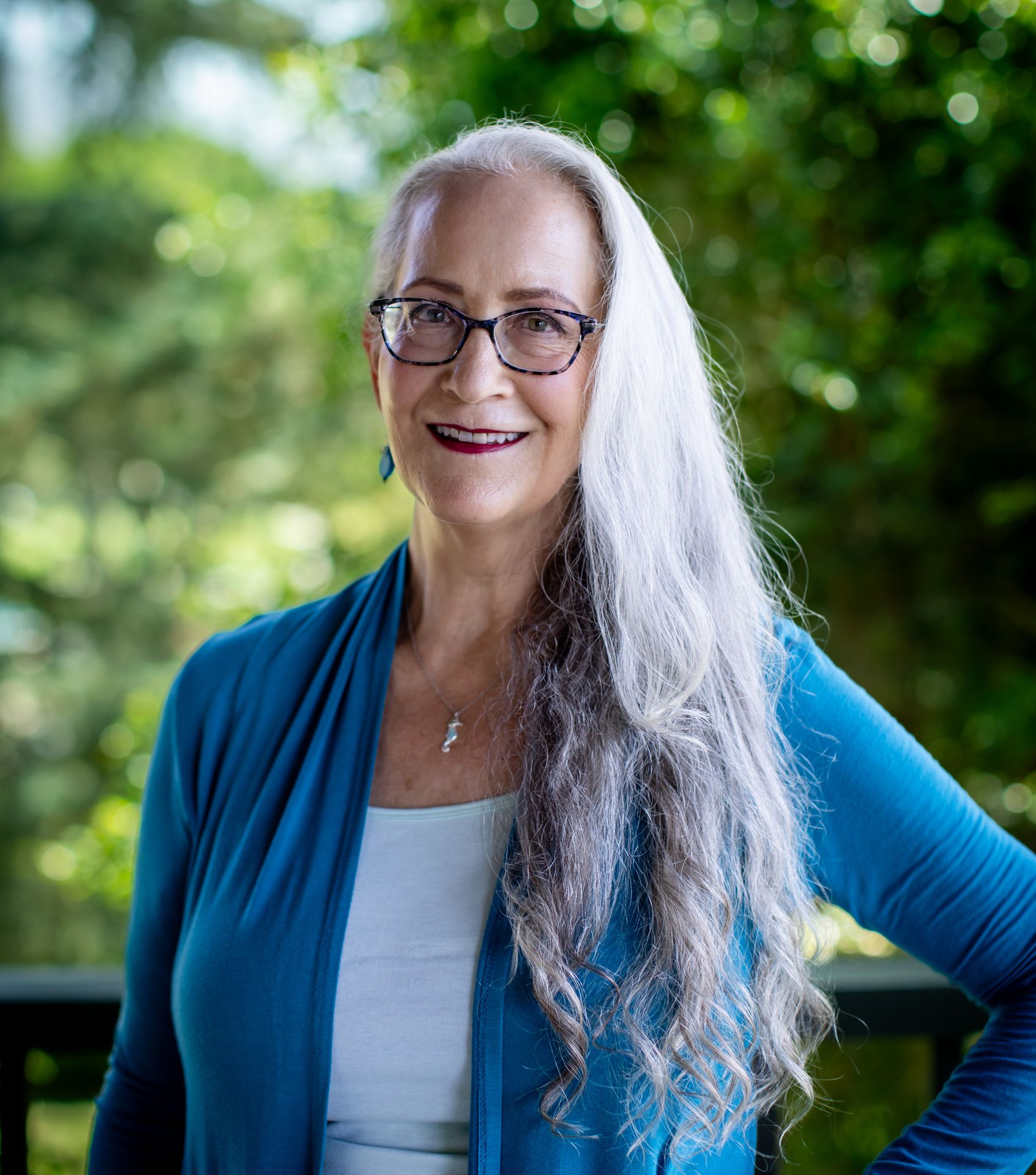 Abby McCarrel, a warm and experienced psychotherapist with long white hair and glasses, stands outdoors with her hand on her hip, surrounded by lush greenery. Her calm, grounded presence reflects her work supporting working parents facing stress, offering online parenting therapy rooted in brain-based, trauma-informed care to help families restore balance and connection at home.