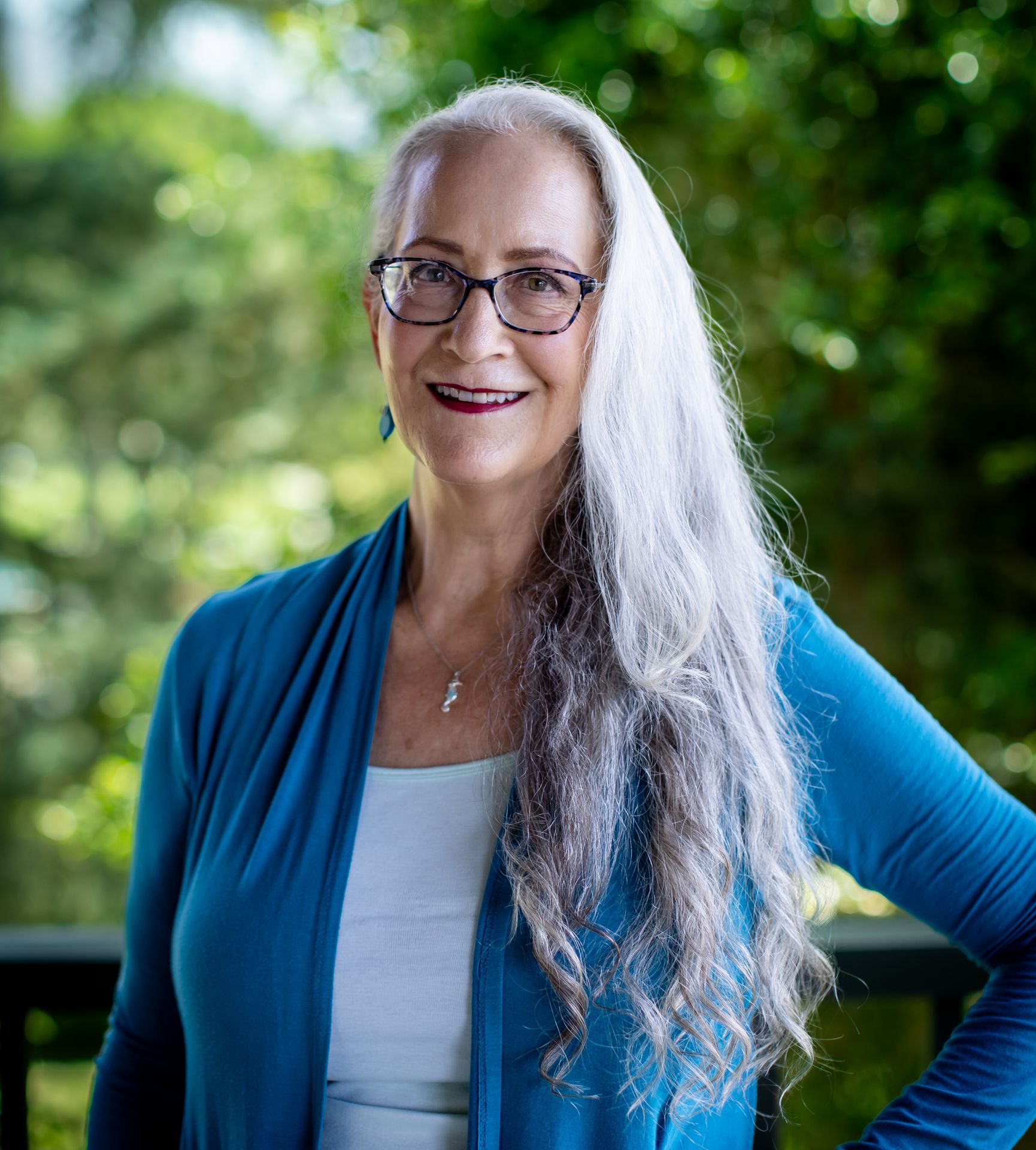 Abby McCarrel, a warm and experienced psychotherapist with long white hair and glasses, stands outdoors with her hand on her hip, surrounded by lush greenery. Her steady, compassionate presence reflects her expertise in parenting neurodiverse and ADHD children, offering brain-based, trauma-informed support to help parents navigate challenges, reduce conflict, and build stronger, more connected relationships with their uniquely wired kids.