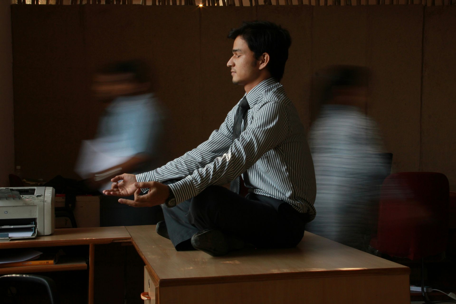 man wearing office work clothes sitting on a desk meditating, while two blurry figures walk by. Each are oblivious to one another