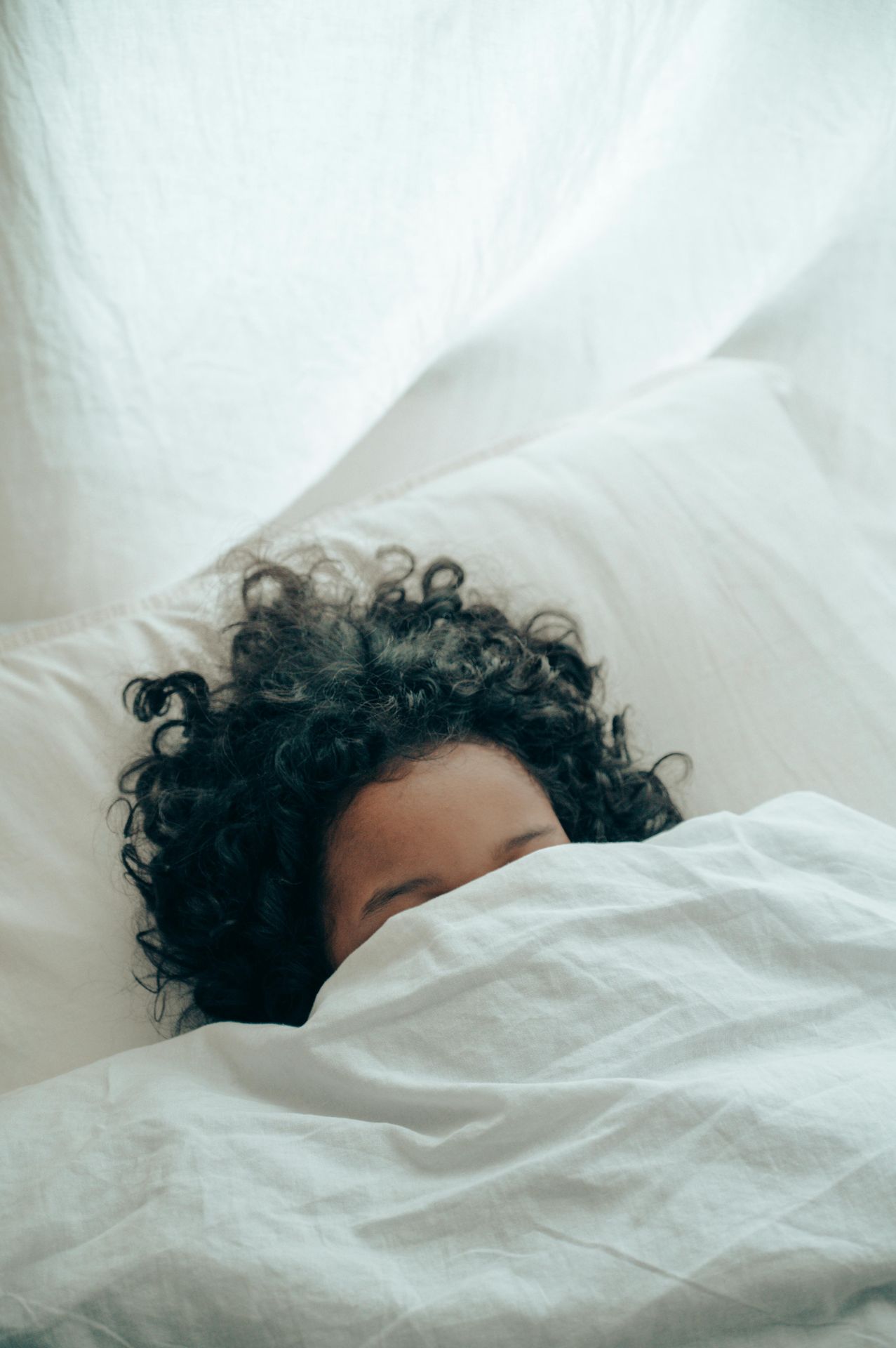 woman with brown curly hair sleeping under a white sheet. perinatal and postpartum sleep issues mental health alaska