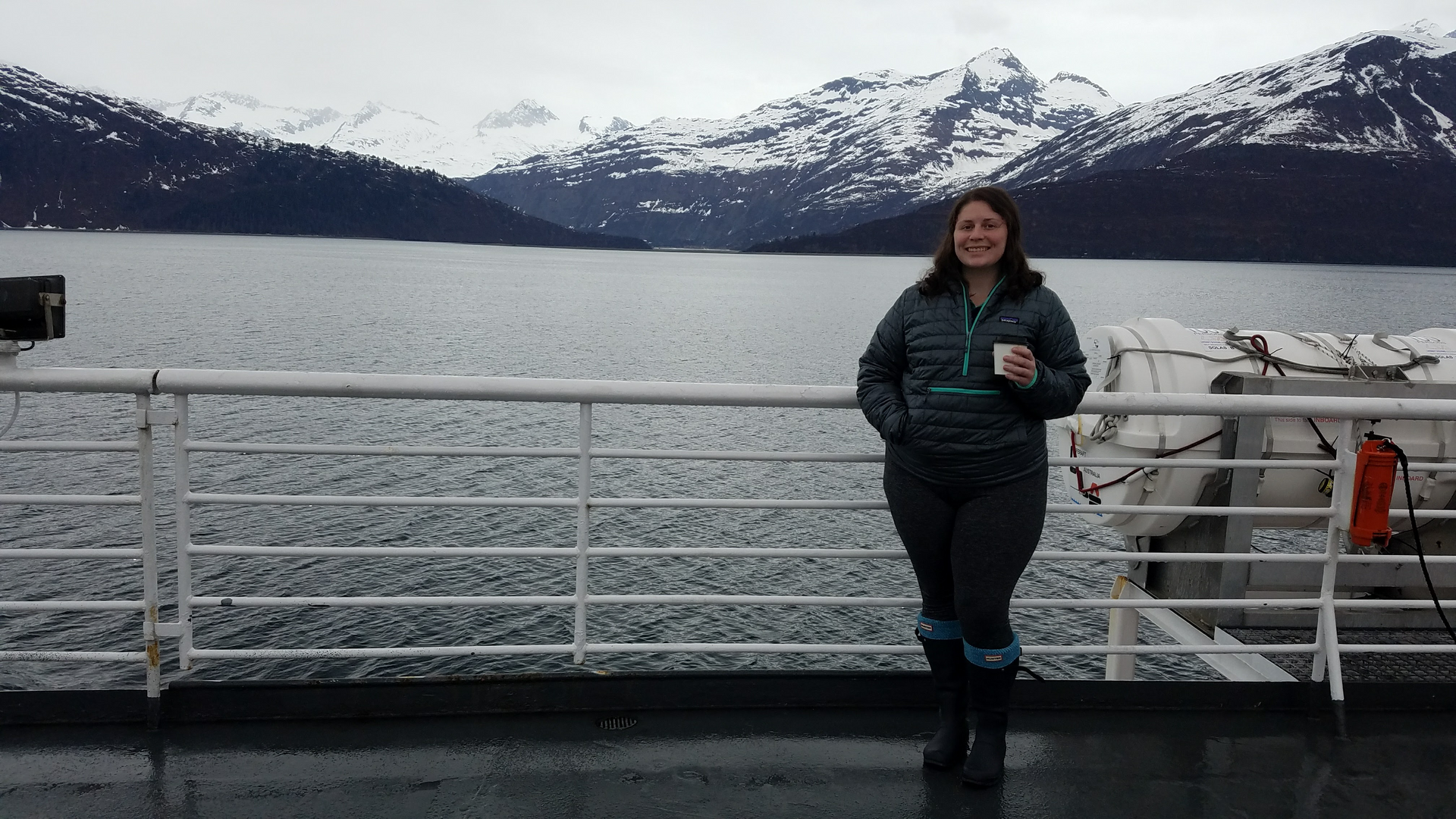 Nicole, a white woman, standing on an Alaska Ferry deck smiling and holding a white cup with mountains in the background