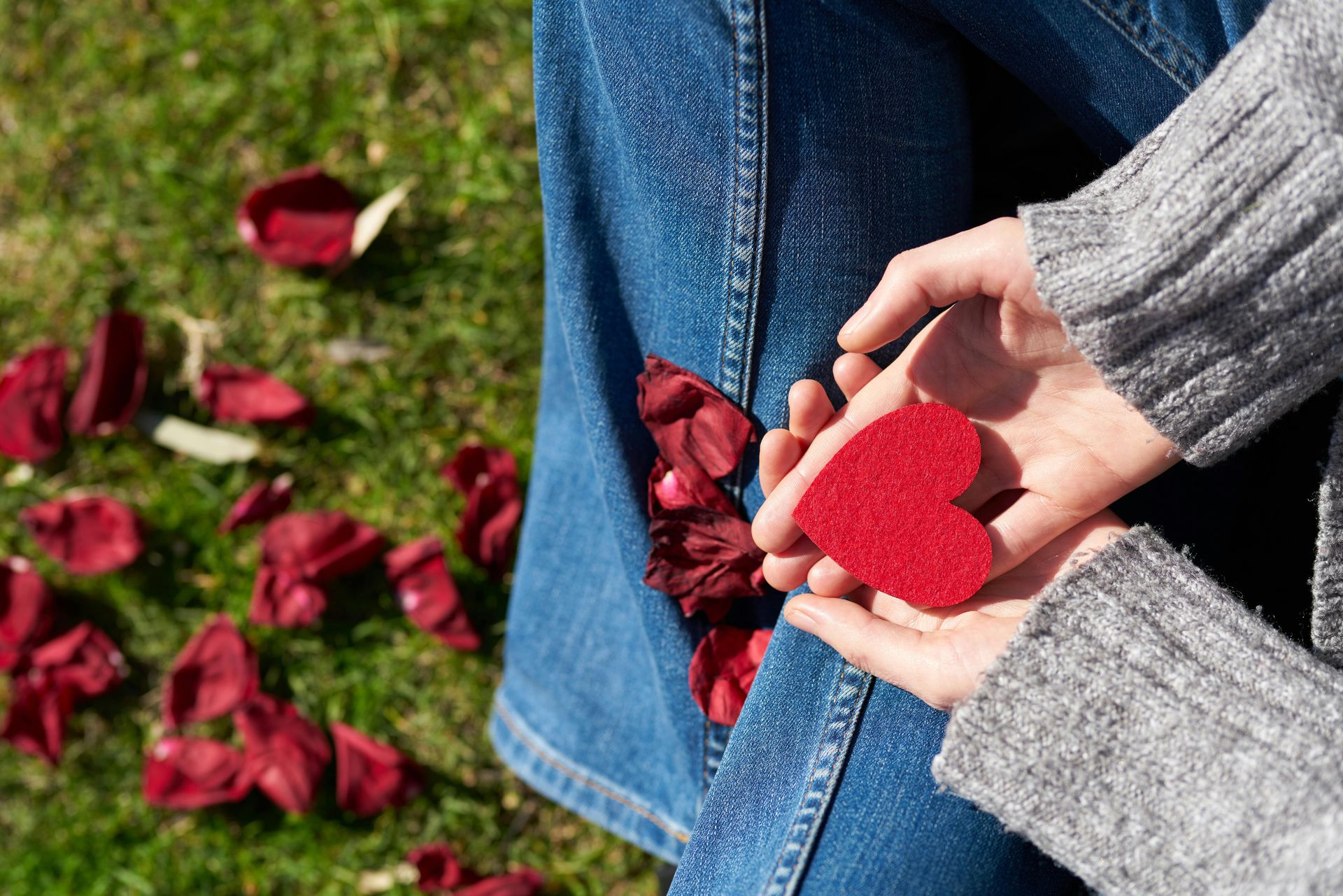 white hands holding a red cut out valentine sitting on the grass with rose petals tips to manage holiday stress from therapist