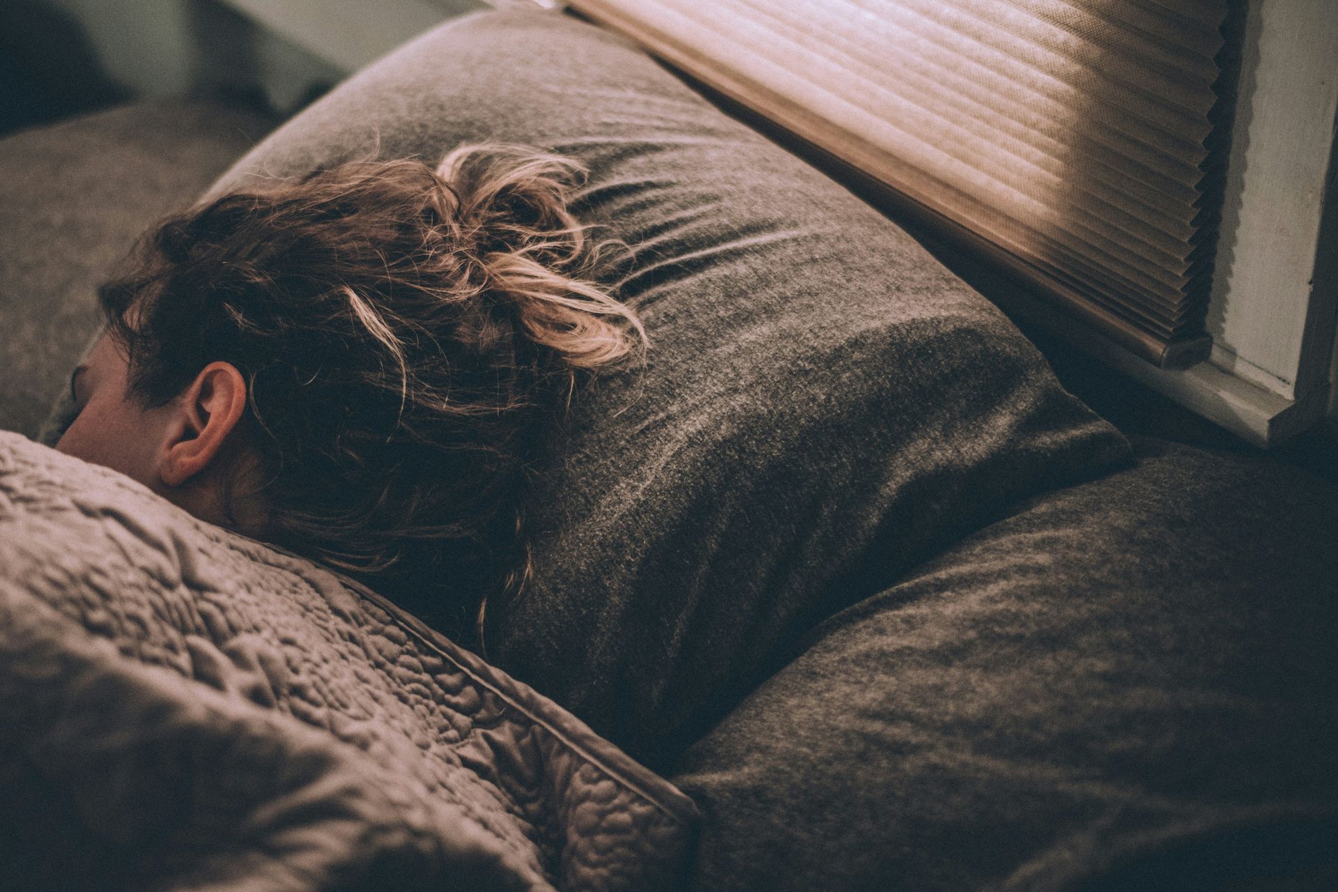 Femme person laying in bed, most of her face is covered by a blanket. Connection between sleep and mental health