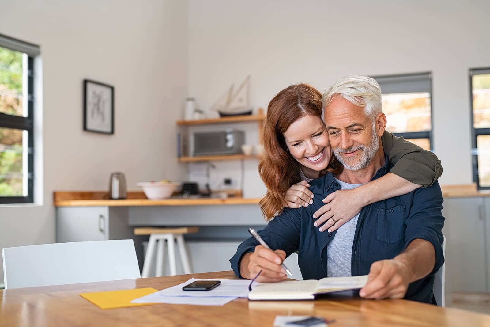 A woman hugging her partner from behind as he writes in a notebook | men’s issues in san francisco, ca | men's therapy san francisco, ca | counseling for men san francisco | 94608 | 94609 | 94061
