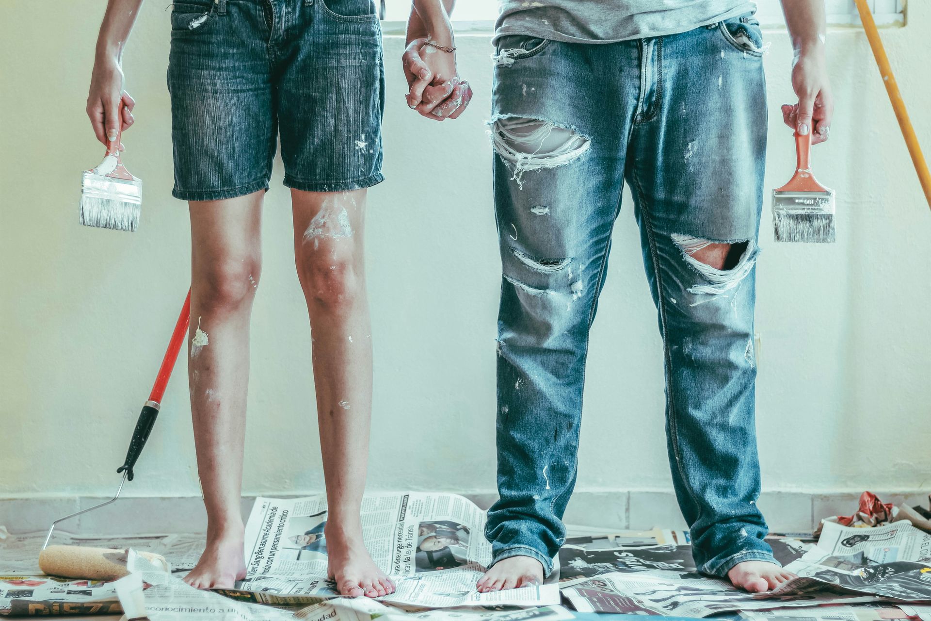 the lower half of th bodies of  a woman and a man painting with paint splattered clothes and painting supplies 