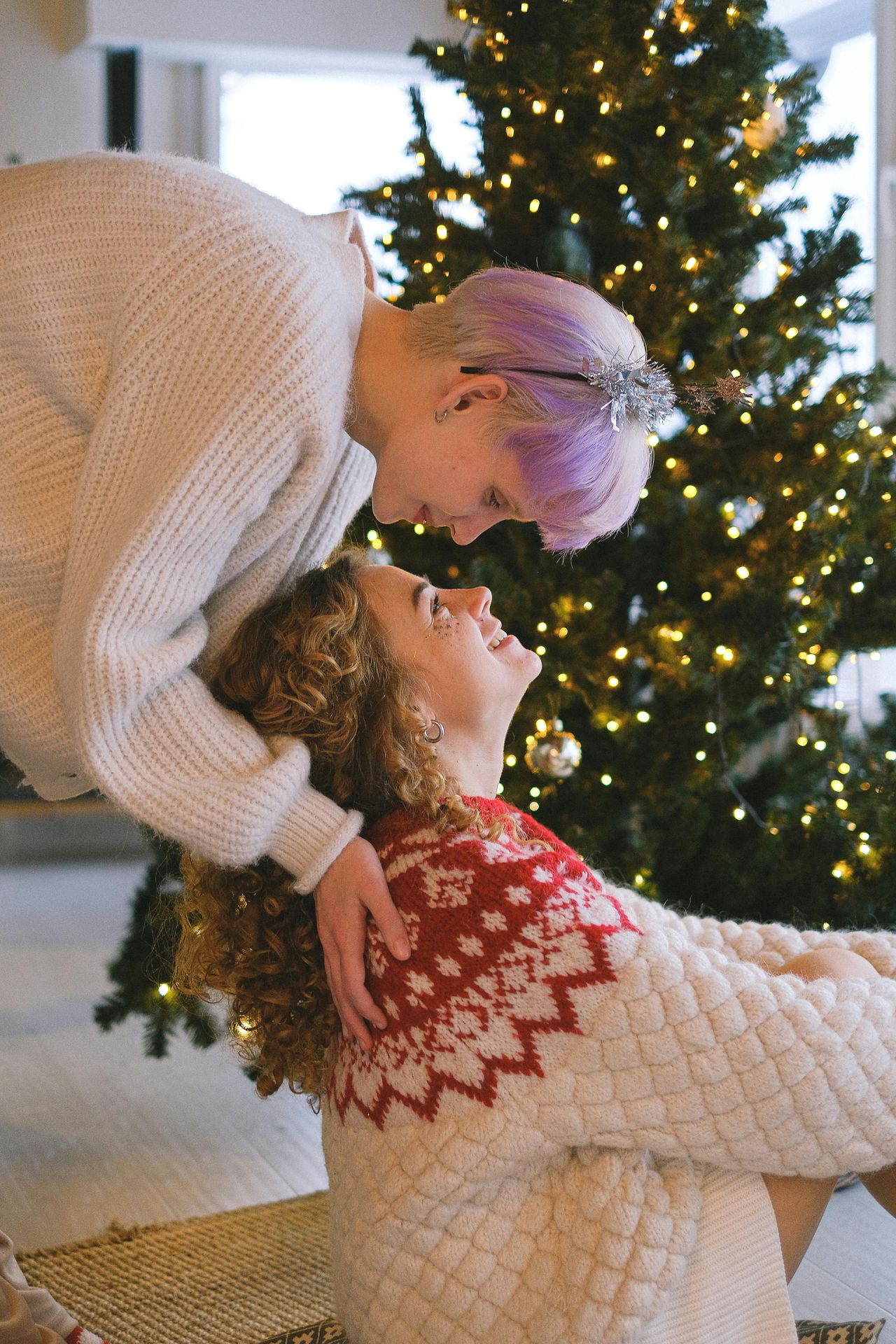 lesbian couple in front of a christmas tree one sitting on the floor the other standing over her both looking into each other's eyes, uplift and connect counseling, Katheryn Barton