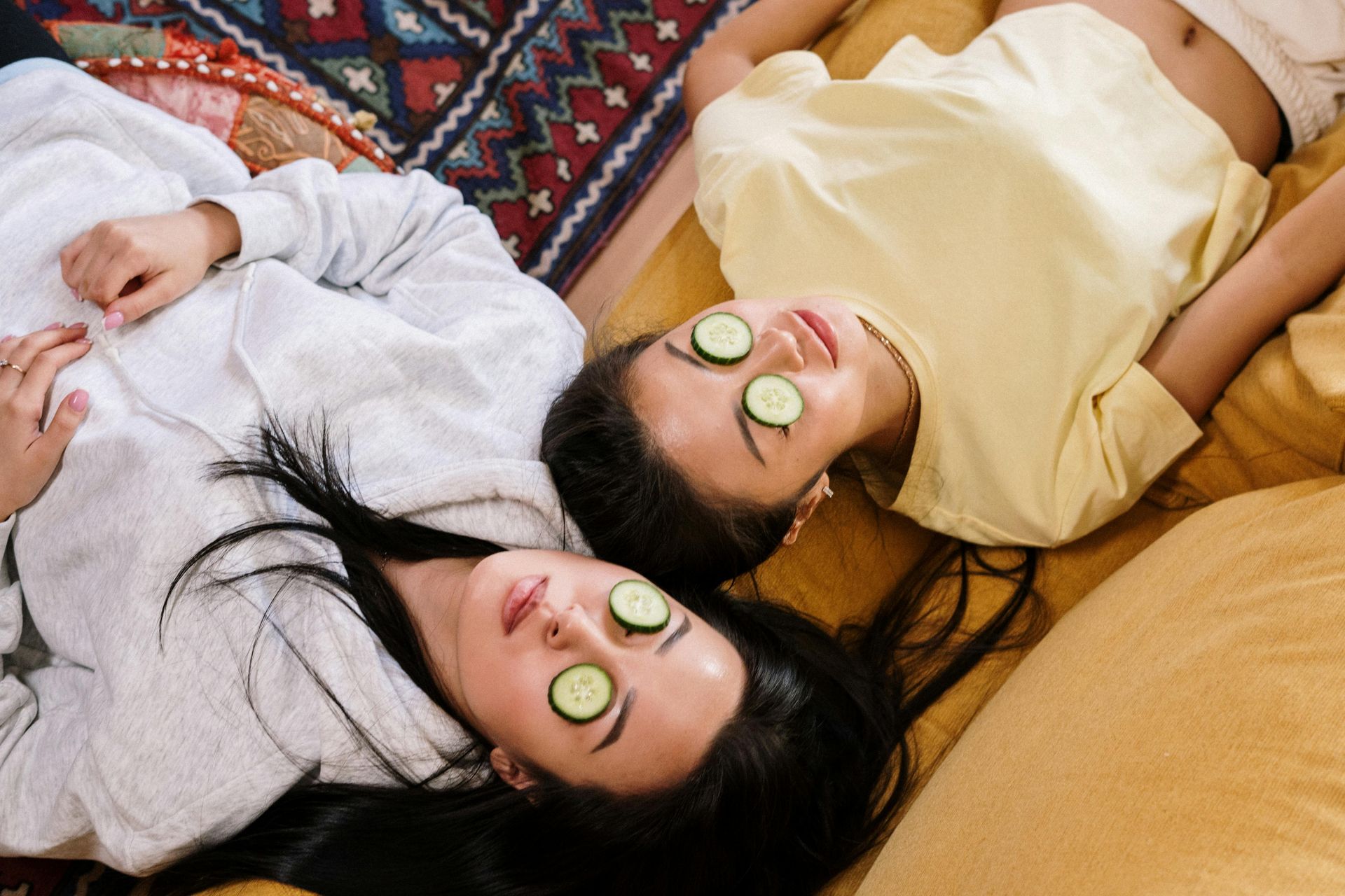female couple lying down with their heads next to each other with cucumbers covering their eyes