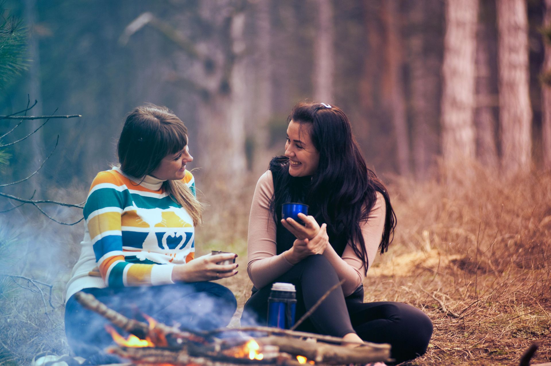 female lesbian couple sitting int he woods by a fire chatting both are holding small cups