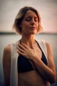 A woman standing near the water with her hand near her neck and her eyes closed. She is practicing self-care and self-compassion that she learned through therapy in Katy, TX 77494