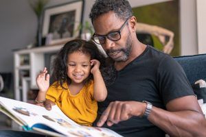 A loving father of African descent sits on the couch at home and reads a storybook to his preschool age daughter. The child is sitting on her father's lap and is smiling while looking at the book, How to Motivate Kids in a Healthy Way blog, counseling center at cinco ranch, counseling in katy texas, richmond texas