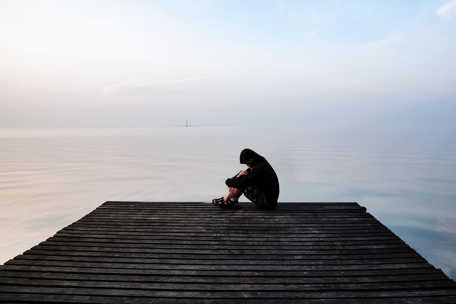 A person in a dark hoodie sits alone on the edge of a wooden pier, hugging their knees and looking down, with calm water and a pale sky in the background. This could represent the isolation of trauma that trauma treatment in Palm Beach County, FL can help you overcome. Learn more about complex PTSD treatment in Palm Beach, FL by contacting a complex PTSD therapist in Palm Beach, FL today. 