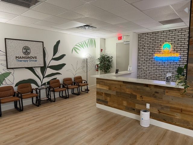 The fully completed reception area of Mangrove Therapy Group's new office, featuring a reclaimed wood reception desk with the glowing practice neon sign, a row of warm leather chairs, a tropical botanical mural, and lush potted plants throughout. This welcoming space sets the tone for the entire experience at this leading Delray Beach therapy practice.
