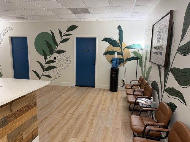 The waiting area of the new Mangrove Therapy Group office, featuring bold Santorini Blue therapy room doors, a sweeping tropical botanical mural, warm wood flooring, leather waiting chairs, and the practice logo displayed on the wall. This thoughtfully designed space reflects the commitment of this EMDR therapy in Delray Beach, FL practice to creating a calm environment.