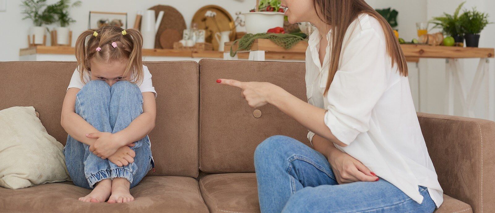 A young girl sits on a couch with her knees pulled to her chest, looking down with a sad expression while an adult woman, possibly her mother, points at her. This could represent past childhood emotional abuse that a complex PTSD therapist in Palm Beach, FL can help offer support with addressing. Learn more about trauma therapy in Delray Beach, FL and how trauma treatment in Palm Beach County, FL can help.