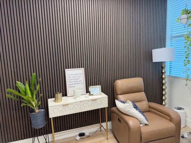 A beautifully finished therapy office corner featuring dark wood acoustic wall panels, a warm leather recliner, a white console table with an inspirational framed quote, a potted plant, and soft lamp lighting. This inviting, carefully curated space is where clients come to do the deep work of trauma therapy in Delray Beach, FL — in an environment designed to feel safe, calm, and grounding from the moment they sit down. For those seeking EMDR therapy in Delray Beach, FL, this is the kind of thoughtfully designed space where real healing happens.