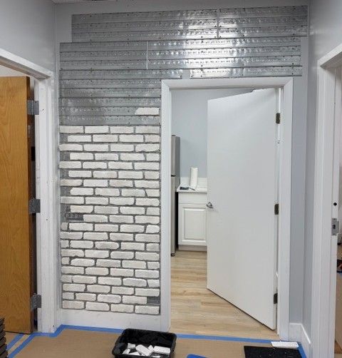A mid-construction view of the hallway area showing white Koni Old Chicago brick veneer being applied to the wall using a metal grid system, with mass-loaded vinyl sheets visible above and a therapy office doorway framed in the background. This image captures the meticulous soundproofing and design work that went into building a space worthy of serious trauma therapy in Delray Beach, FL. For clients of this Delray Beach mental health therapist practice, the result is a confidential, calm, and beautifully finished environment where they can feel truly safe.