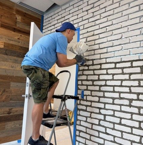 A team member stands on a ladder, carefully applying and shaping grout between white Koni Old Chicago brick tiles on the accent wall, with reclaimed wood paneling visible on the adjacent wall behind a blue door. This image captures one of the most labor-intensive phases of the office build-out for this trusted therapist in Delray Beach, FL.