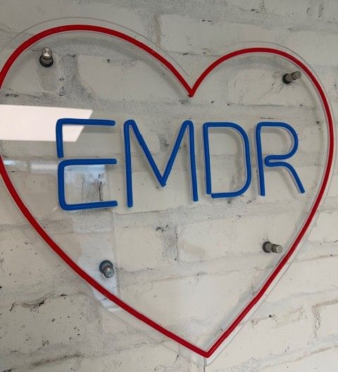 A custom neon sign reading "EMDR" in blue letters inside a red heart shape, mounted on the white brick accent wall at Mangrove Therapy Group's new office. This sign is a nod to the practice's deep expertise in EMDR therapy in Delray Beach, FL.