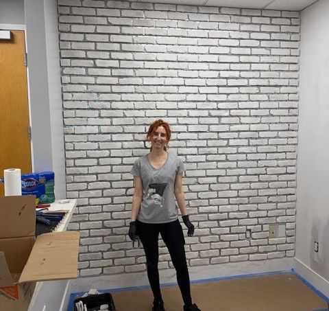 A smiling team member stands proudly in front of a freshly completed white Koni Old Chicago brick accent wall, wearing work gloves and a casual outfit after a long day of hands-on construction work. This wall represents one of the most labor-intensive elements of the build-out at Mangrove Therapy Group, one of the most trusted names in trauma therapy in Delray Beach, FL. The finished result is a striking, textured feature that adds warmth and character to the new home of this EMDR therapy in Delray Beach, FL practice.