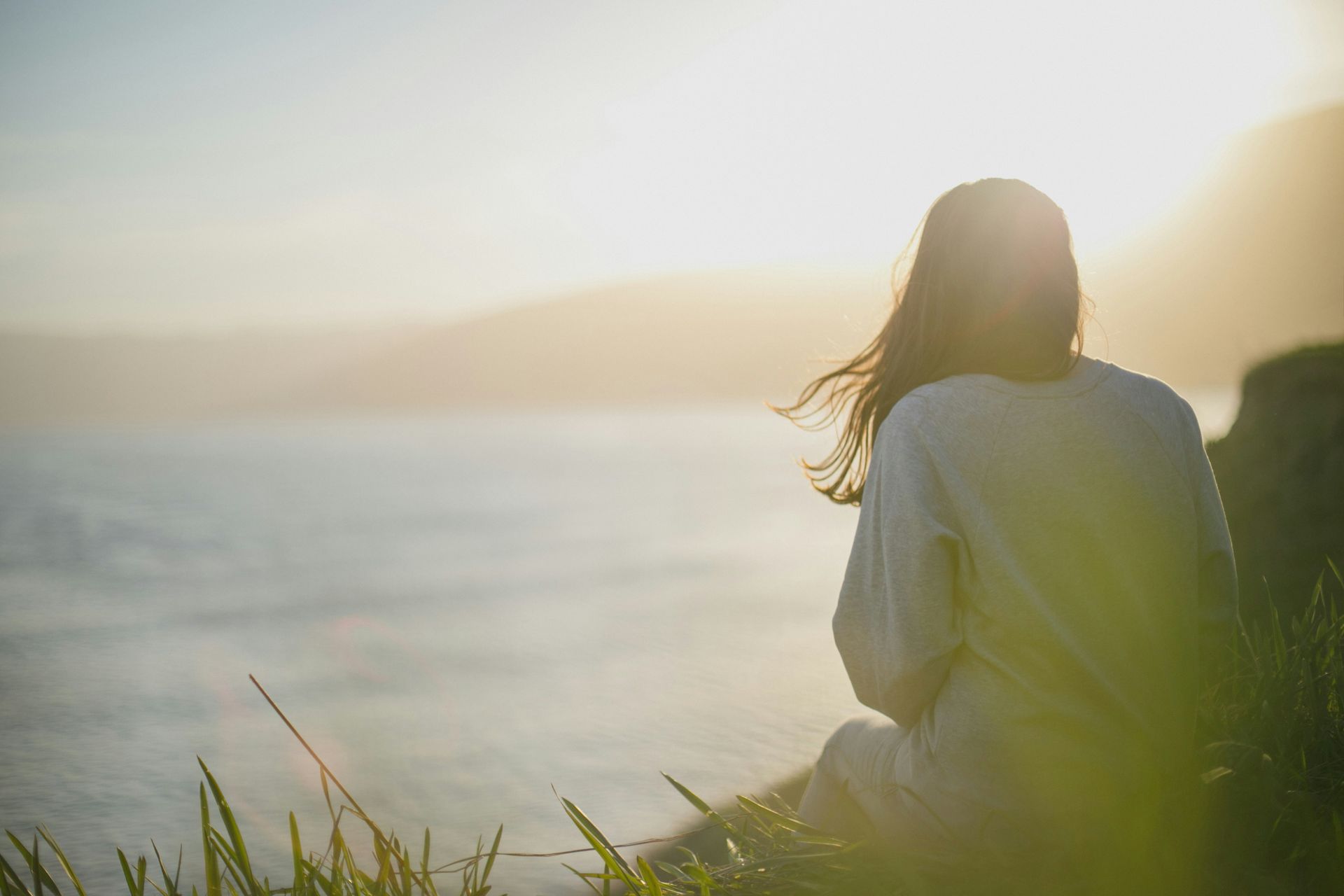 A woman sits peacefully on a grassy cliffside overlooking the ocean at sunrise, representing moments of reflection during trauma therapy in Delray Beach, FL. The calm scene mirrors the emotional clarity often achieved through working with a PTSD therapist in Delray Beach, FL or engaging in trauma treatment in Palm Beach County, FL.