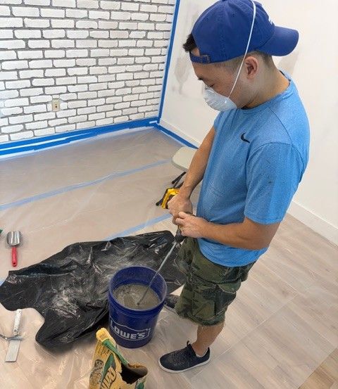 A team member wearing a blue cap and mask carefully mixes grout in a bucket, preparing to apply mortar between the white brick veneer tiles being installed on the walls of the new Mangrove Therapy Group office. This meticulous, hands-on process reflects the level of dedication behind building one of the most thoughtfully designed spaces for trauma therapy in Delray Beach, FL.