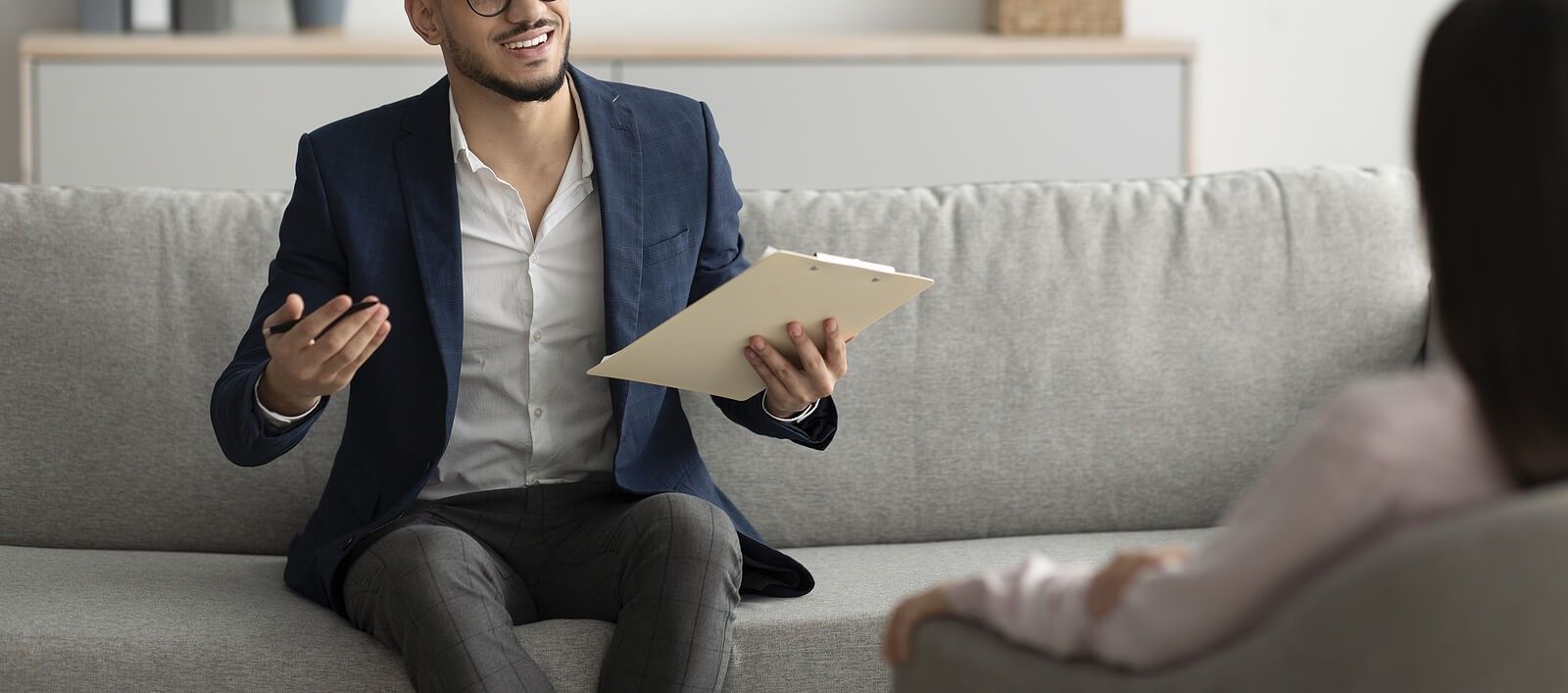A man with glasses, wearing a navy blazer and gray pants, sits on a gray couch while holding a clipboard. He is smiling and gesturing warmly as he talks to a client sitting across from him. This could represent how an EMDR therapist in Palm Beach County, FL can offer support with reprocessing memories. Search for EMDR therapy in Boca Raton, FL and how EMDR group therapy in Palm Beach County, FL can help today. 33431 | 33432 | 33486