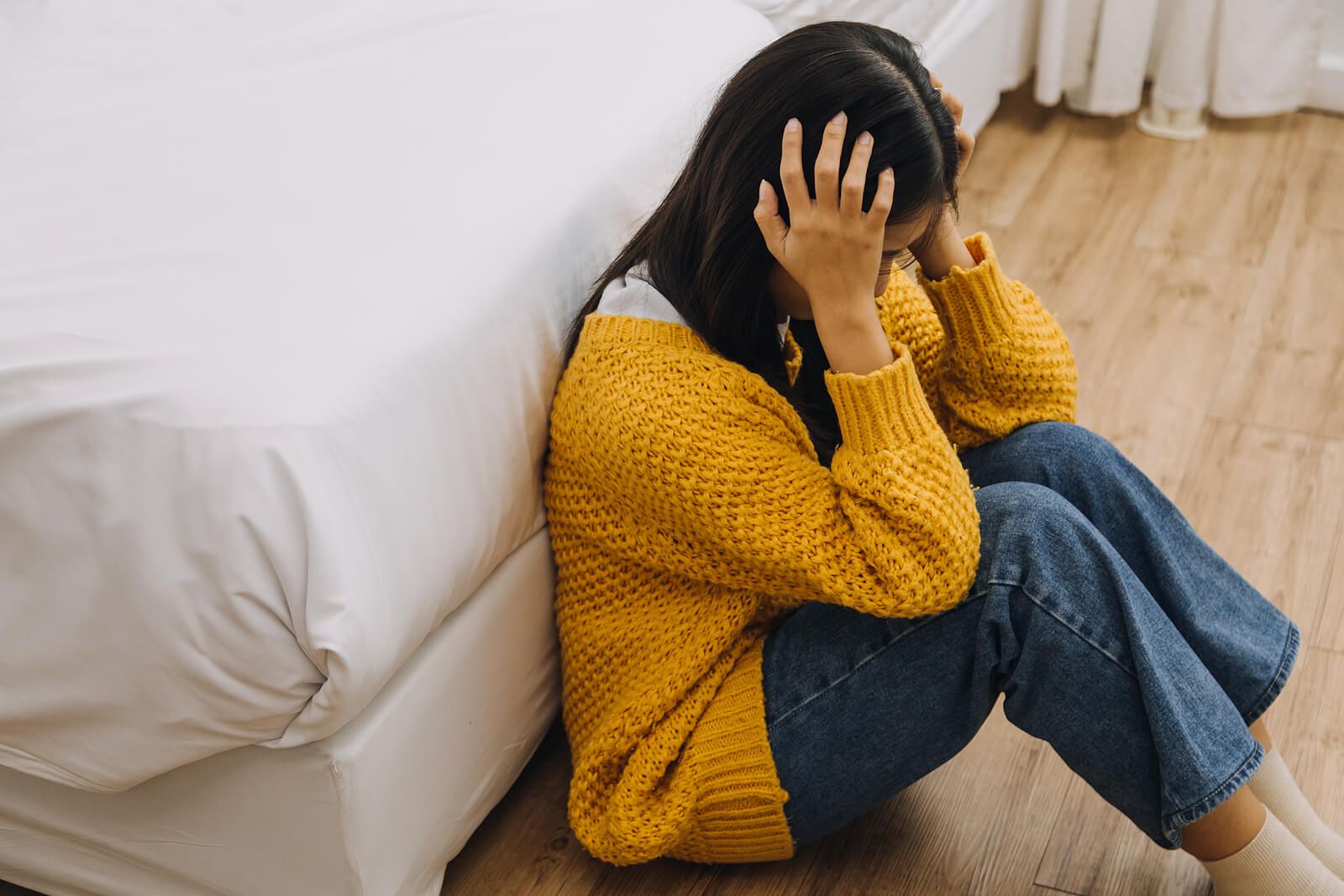 A young woman in a yellow sweater sits on the floor with her back against a bed, head in her hands in a moment of quiet overwhelm. This image reflects the invisible, cumulative weight that brings many people to seek complex PTSD treatment in Palm Beach, FL — the kind of pain that doesn't always have one clear cause but shows up in the body and in everyday moments. A complex PTSD therapist in Palm Beach, FL understands that this kind of suffering is real, valid, and treatable.