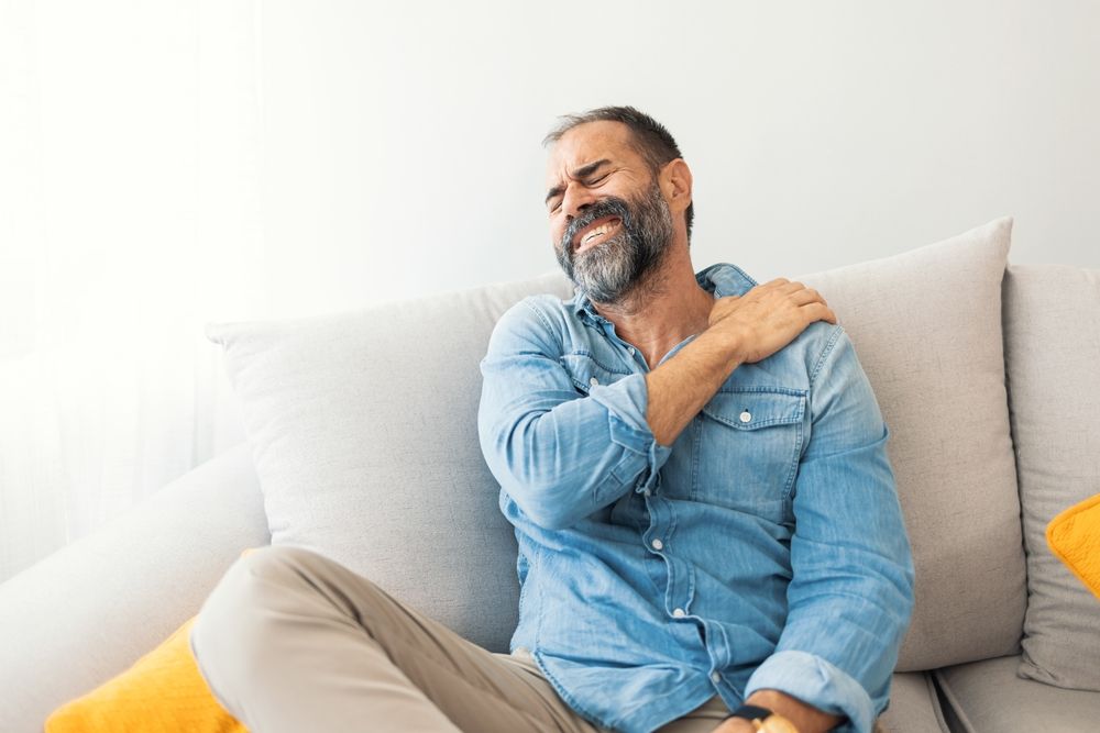 Person wincing and holding their shoulder while sitting on a couch, illustrating chronic pain and the mind-body stress cycle. Image for emdr therapy in palm beach, fl, emdr therapy in delray beach, fl, and support from an emdr therapist in palm beach county, fl.
