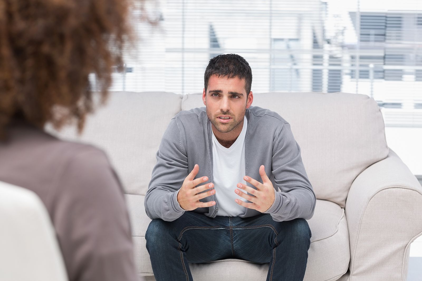 A man in casual clothing sits on a light-colored couch, speaking with a concerned expression to a person whose back is visible in the foreground, indicating a therapy or counseling session.