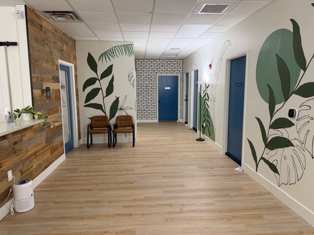 A wide view of the fully completed hallway at Mangrove Therapy Group, lined with Santorini Blue therapy room doors, reclaimed wood paneling, white brick accent walls, and a sweeping tropical mural that stretches the full length of the corridor. This stunning space is the result of months of hands-on work by the team behind one of the most respected practices for trauma therapy in Delray Beach, FL.