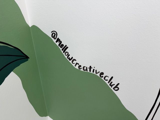 A close-up of a section of the hallway tropical mural at Mangrove Therapy Group, featuring lush green botanical leaves painted directly on the wall alongside the artist's Instagram handle, @mellowcreativeclub. This custom mural was commissioned to bring warmth, life, and a sense of calm to the hallway of this EMDR therapy in Delray Beach, FL practice.
