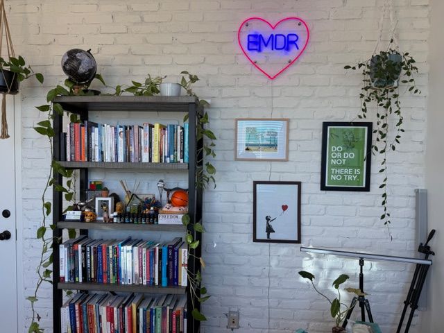 A fully decorated therapy office wall featuring the glowing EMDR neon heart sign, a tall bookshelf filled with clinical texts and personal touches, trailing green plants, and framed motivational artwork against the white brick backdrop. This warm, lived-in space reflects the personality and clinical depth of this trusted therapist in Delray Beach, FL — a room designed to feel both professional and deeply human.