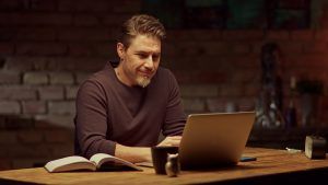 A man sits at his kitchen table in front of his laptop as he smiles. This could represent meeting with an online therapist in Florida. Contact us to learn about online EMDR therapy, online therapy, and other services.
