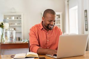 A man smiles as he types on his laptop resting on the kitchen counter. Online therapy in Florida can provide support from the comfort of home. Contact an online therapist to learn more about online anxiety treatment, online trauma therapy, and other services.