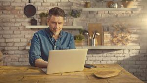 A calm, content man smiles at his kitchen counter as he looks at something on his laptop. This could symbolize the peace of mind that comes from online EMDR therapy in Florida. Contact an EMDR therapist in Tampa, FL to learn how online emdr in Florida can support you!